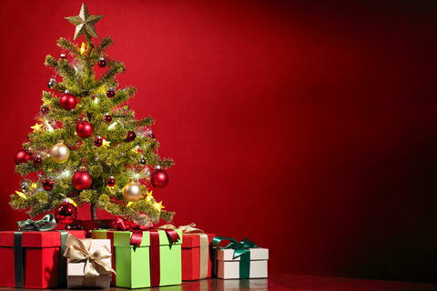 Red background with a decorated christmas tree on the left hand side with christmas presents on the floor under it.